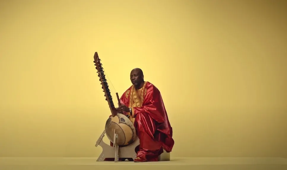 A musician in traditional attire, seated, plays a kora
