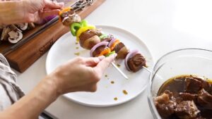 A person assembles shish kabobs by threading marinated meat and vegetables onto metal skewers over a white plate
