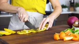 A person slices yellow bell pepper on a wooden cutting board with other chopped vegetables nearby