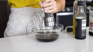 A person grinds black pepper into a glass bowl of marinade next to bottles of oil and balsamic vinegar