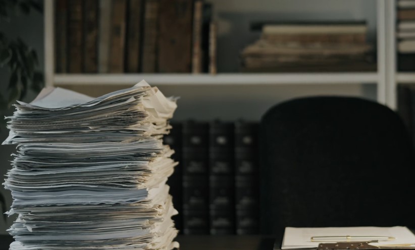 A stack of papers rests on a desk in front of a bookcase filled with books