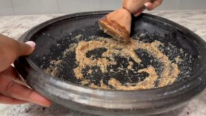 A person grinding ginger, onion, and spices into a paste using a traditional Ghanaian grinding bowl and wooden grinder