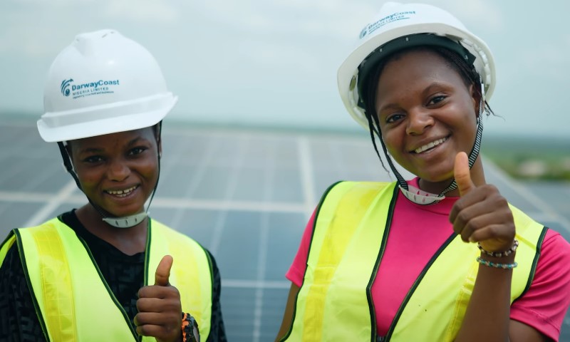 Two women in hard hats and safety vests stand beside a solar panel in Nigeria