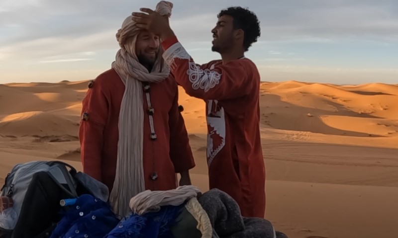 Local guide expertly wraps a turban on a traveler against a backdrop of towering orange sand dunes at sunset