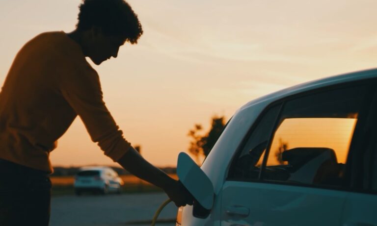 A man charges his electric car at a charging station during a vibrant sunset