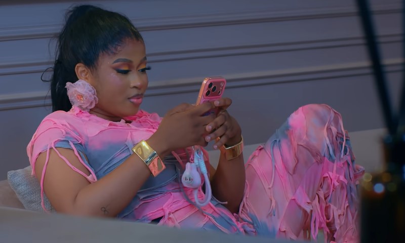 Imisi Ayanwale in a pink shredded outfit relaxes on a couch, focused on her phone