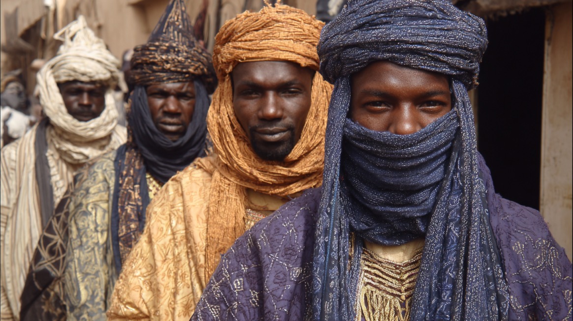 Group of men wearing traditional Hausa attire and wrapped turbans standing in a village setting