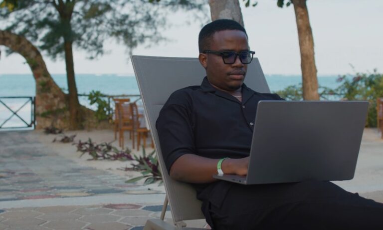 A person in sunglasses sits on a lounge chair outdoors, working on a laptop
