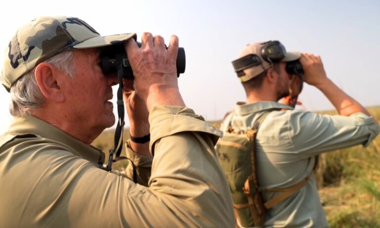 Two people in camouflage hats and khaki shirts use binoculars to observe a wildlife area