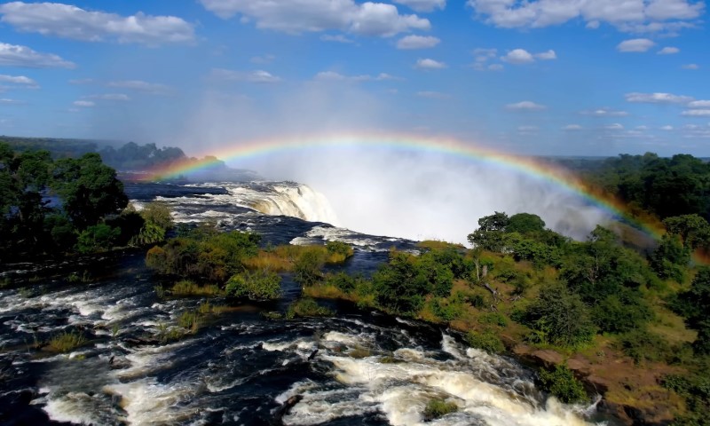 Majestic Victoria Falls cascades with mist rising, creating a vibrant rainbow under a bright blue sky