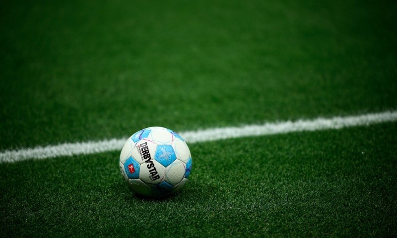 A blue and white soccer ball rests on a vibrant green field, positioned near a white sideline. The scene is calm and anticipates upcoming action.
