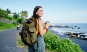 A smiling woman stands near a coastal road, holding a phone and wearing a large backpack