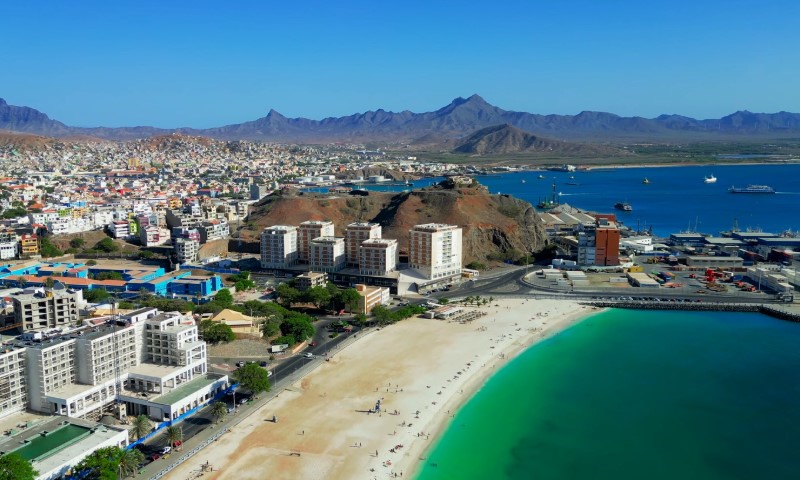 Aerial view of a coastal city: sandy beach with few people, turquoise water, modern buildings, and a busy harbor