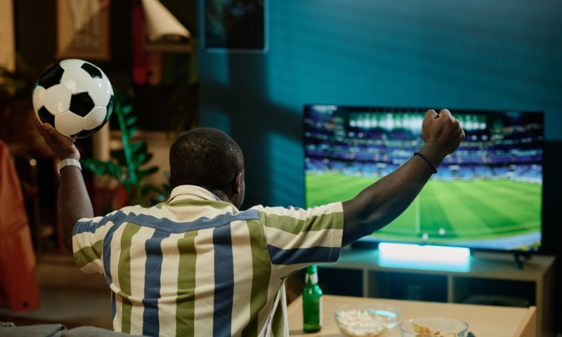 A person in a striped shirt, holding a soccer ball, cheers enthusiastically at a soccer match on TV. A bowl and bottle are on the table nearby.