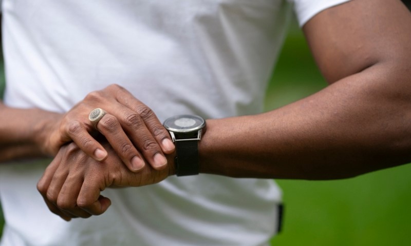 A person wearing a white shirt adjusts a black smartwatch on their wrist. Their other hand displays a ring with a patterned design. The background is blurred green foliage.
