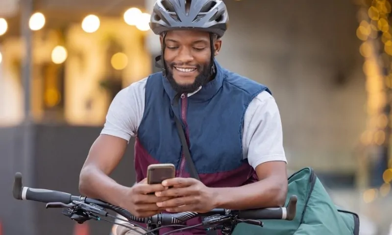 A man in a helmet and vest smiles while checking his phone, sitting on a bike with a delivery bag
