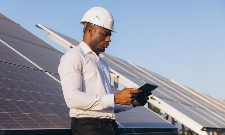 A man in a white hard hat and shirt examines a tablet in front of shiny solar panels