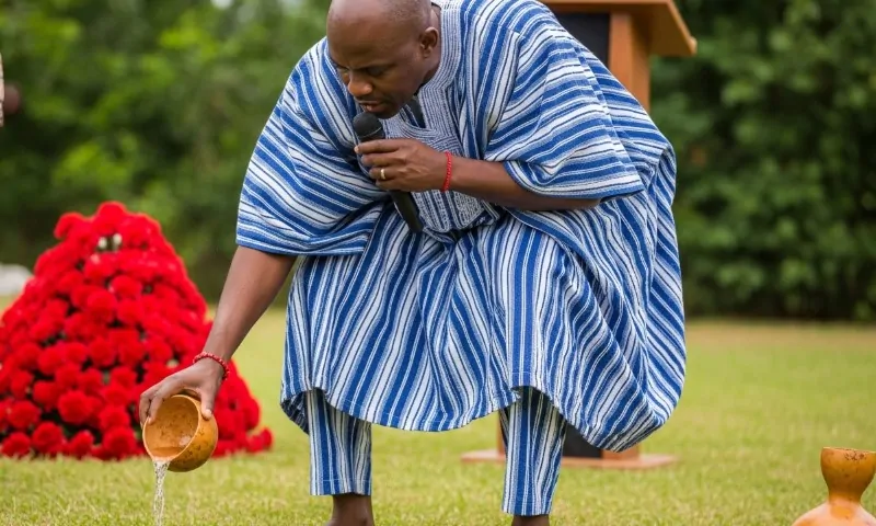 A man in a blue and white striped traditional robe is pouring liquid from a gourd during a ceremonial act