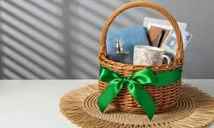 A wicker basket with a green ribbon contains a blue blanket, flowers in a mug, a flask, and notebooks, set on a woven mat