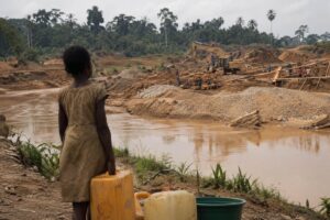 Child holding a water container near a gold mining site and muddy river in rural Ghana.